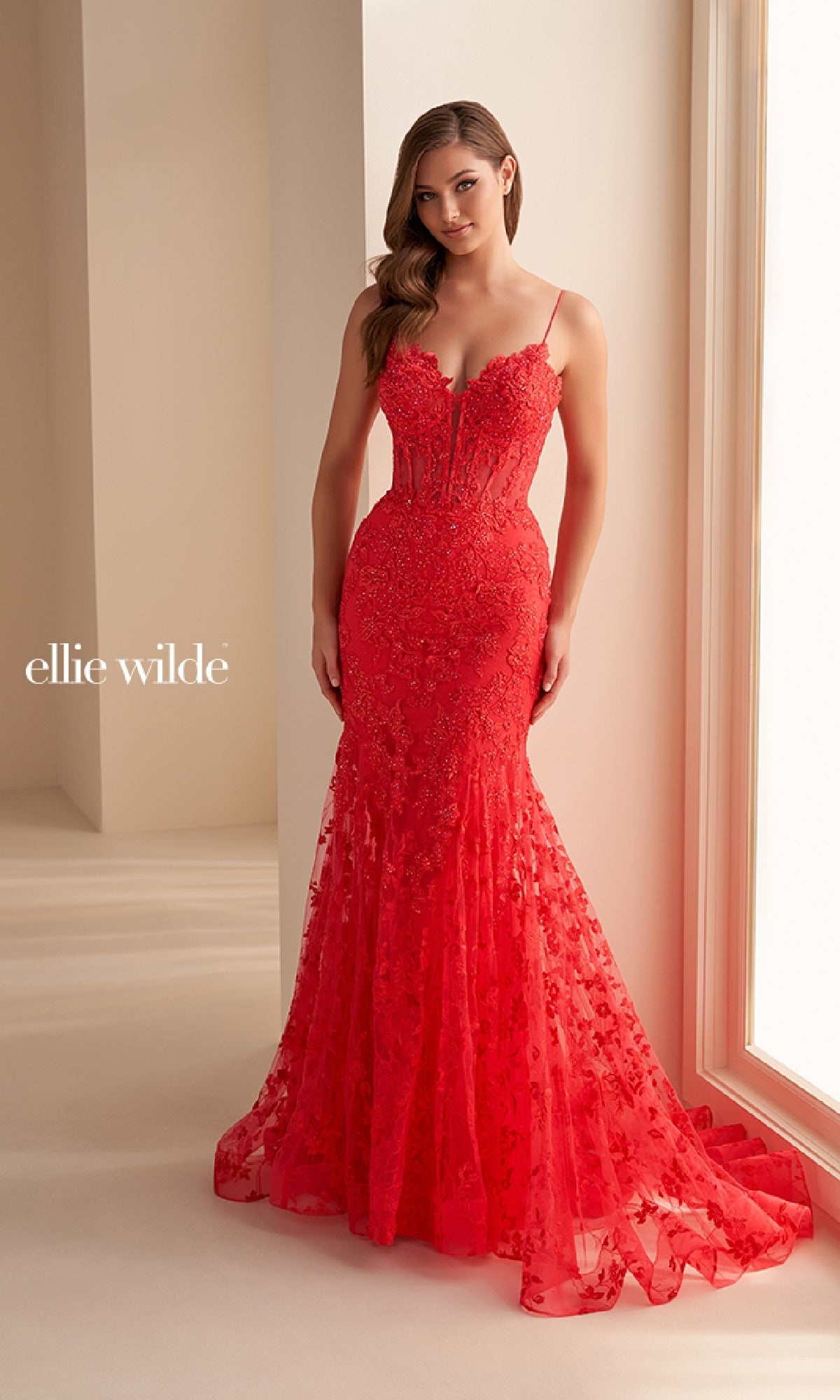 A woman stands indoors near a window wearing the Ellie Wilde EW36008, a fitted red mermaid prom dress with spaghetti straps, intricate embroidery, and a slight train for an elegant, formal look.
